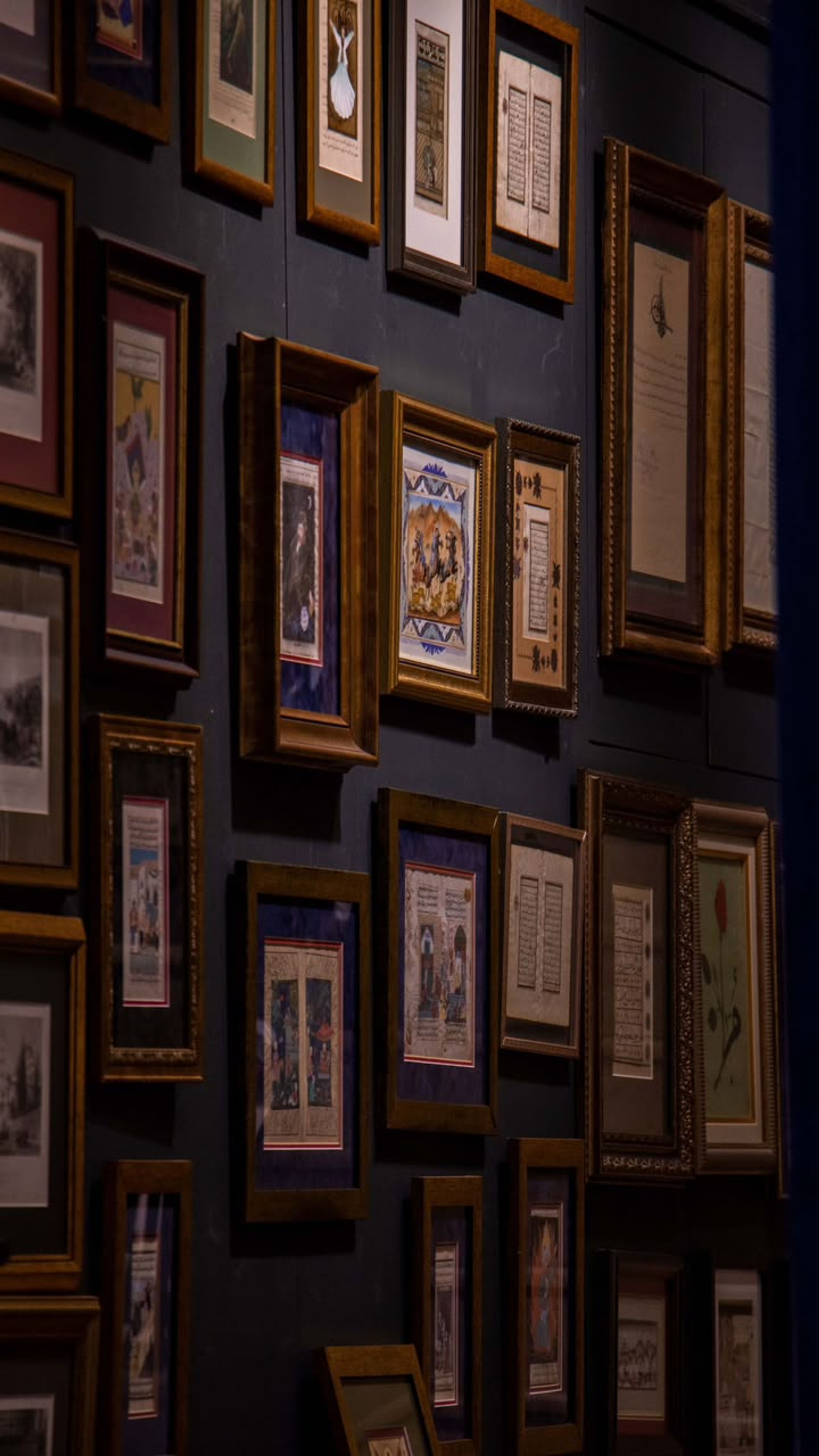 A dark wall hung closely with gilt-framed antique manuscript pages and miniature paintings, lit from the side