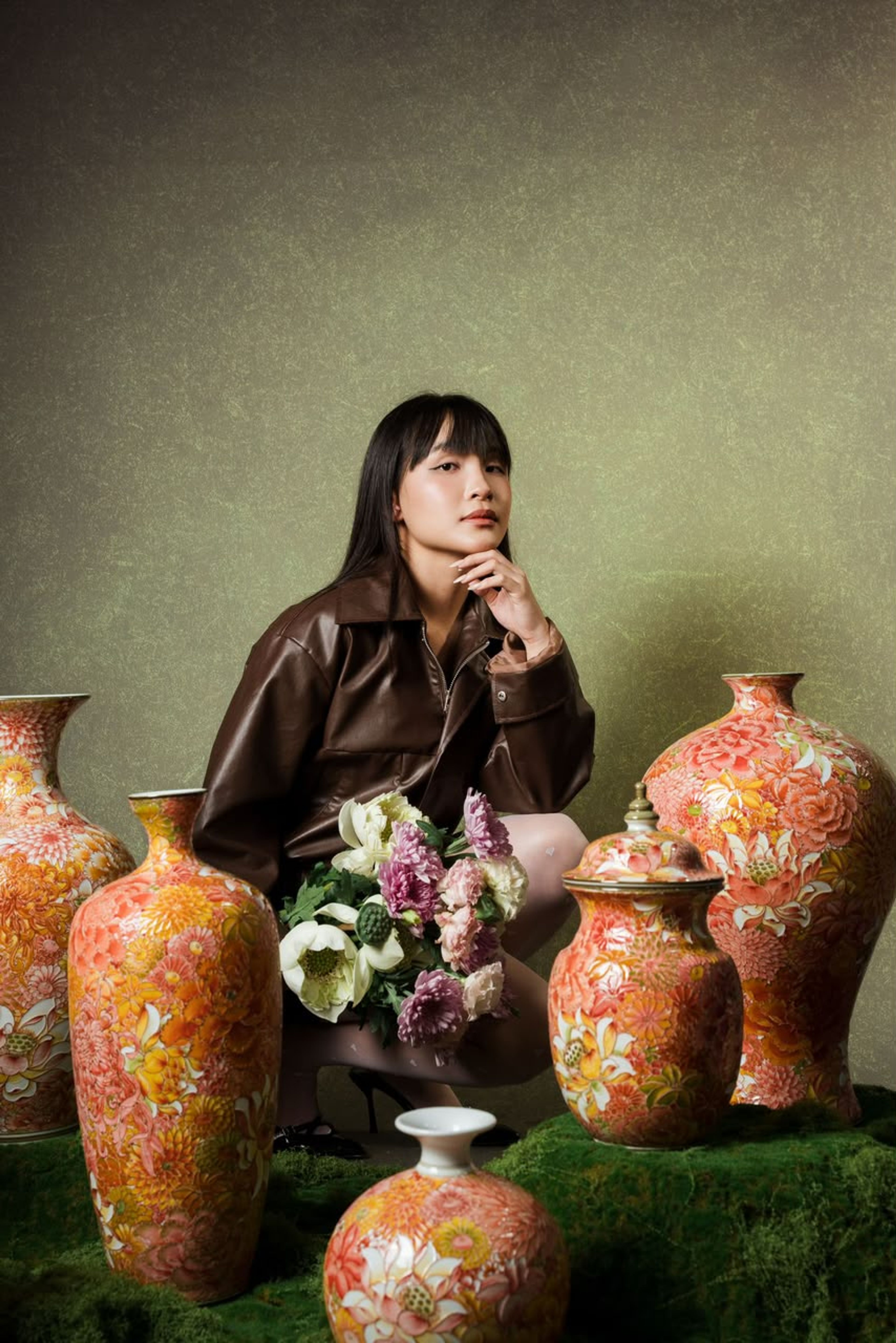 A woman seated among tall orange floral porcelain vases and a bouquet of cream and pink flowers, on a bed of green moss against an olive wall