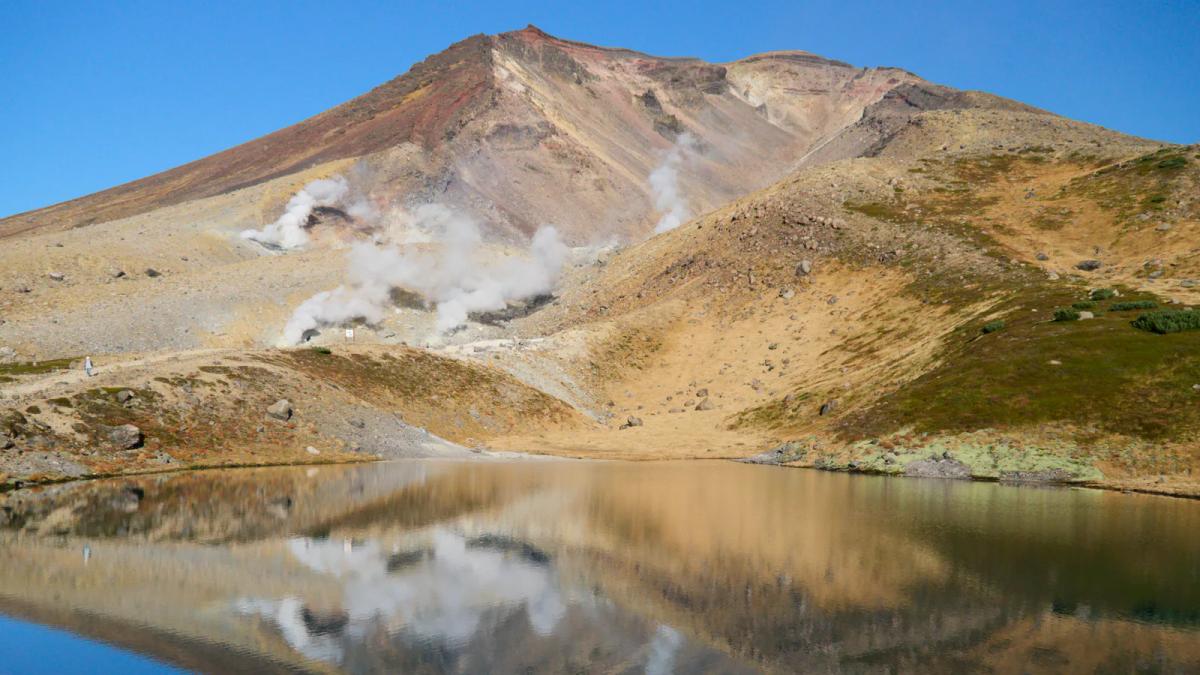 姿見の池倒映著旭岳，山腰的噴氣孔冒著白煙，四周是秋天轉黃的草坡