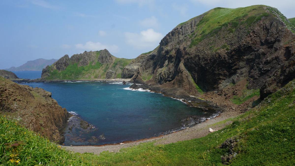 禮文島澄海岬的海灣,藍綠色的海被兩側長滿綠草的岩壁圍住,前方是草坡與礫石灘