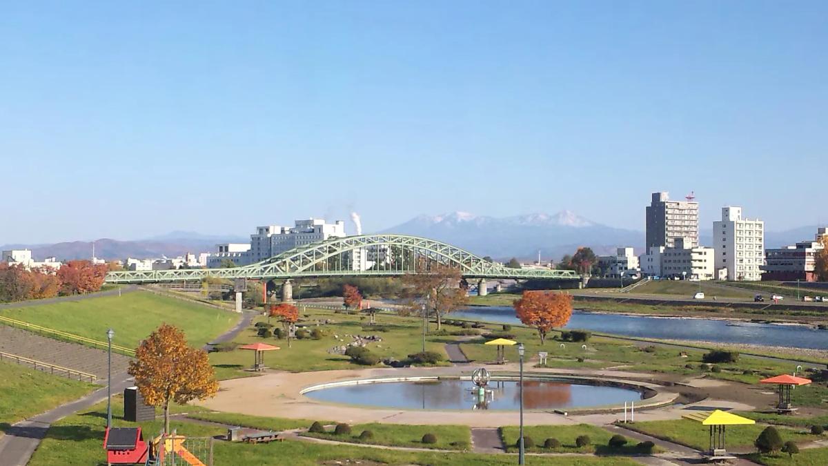石狩川上的綠色鋼造拱橋旭橋，背後是覆雪的大雪山連峰，河岸公園的樹葉轉紅