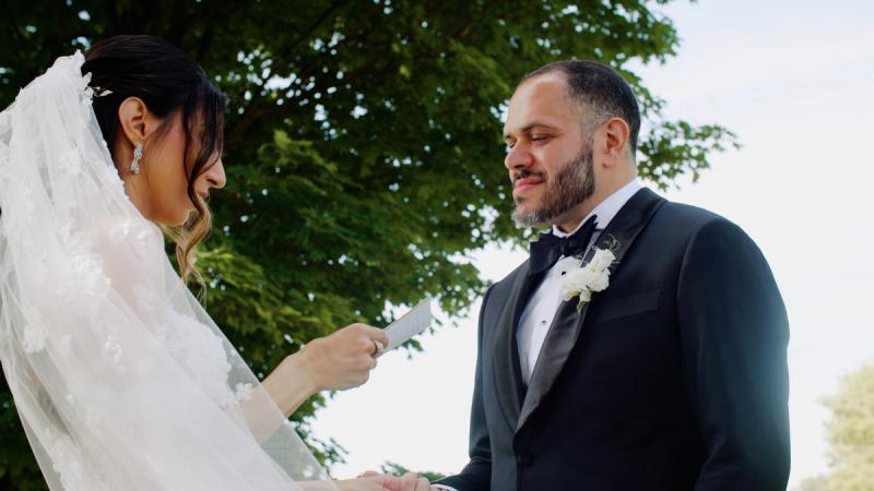 A bride and groom exchange vows beneath a large tree at Springton Manor Farm, surrounded by bridesmaids in sage green gowns and groomsmen in black tuxedos, with white floral arrangements in the foreground.
