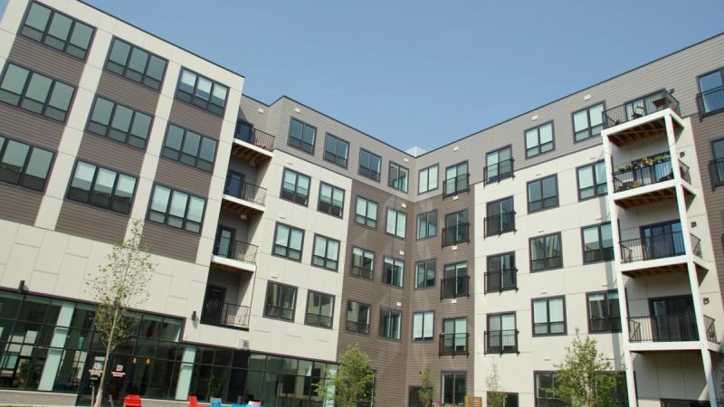 The exterior courtyard facade of Brewer's Block apartments in Pittsburgh, a modern multi-story building with balconies, large windows, and a mix of gray siding and white paneling under a clear blue sky.