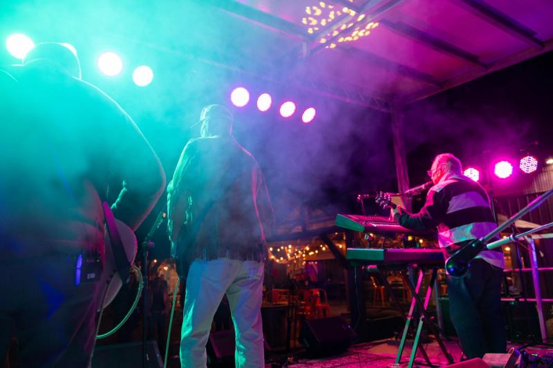 A performer in a leather fringed jacket stands at the front of a stage at Rock Against Cancer, bathed in teal and purple stage lighting with smoke in the air, while a guitarist plays keyboards in the background.