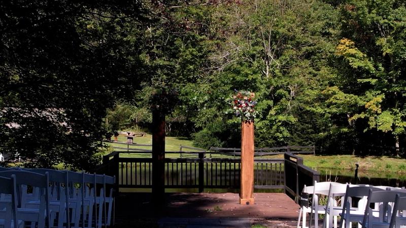Ground-level view of the ceremony space at Chanteclaire Farm, showing a wooden altar post with a floral arrangement, white chairs arranged on either side of a dock, and a pond and treeline in the background.