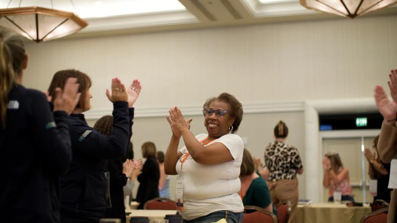 A conference attendee smiles and claps enthusiastically during a standing ovation in a hotel ballroom, surrounded by other participants doing the same.