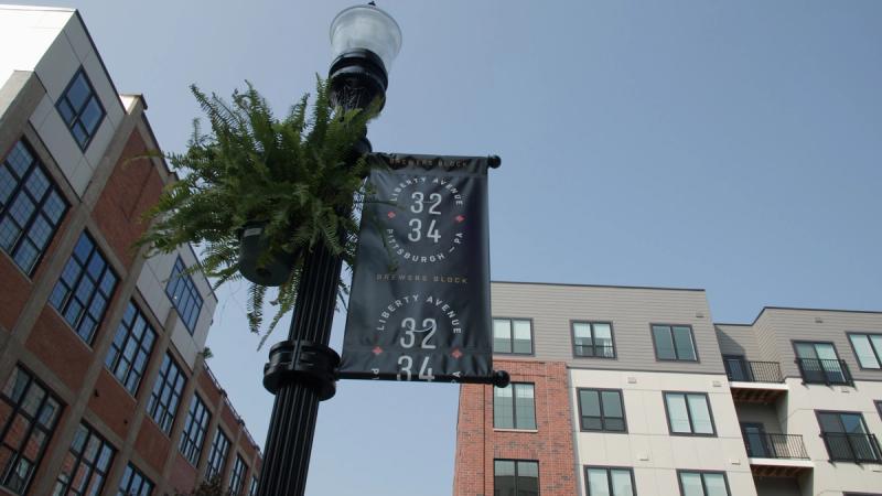 A low-angle shot looking up at a Brewer's Block street banner on a lamp post on Liberty Avenue in Pittsburgh, with the apartment building visible in the background against a blue sky.