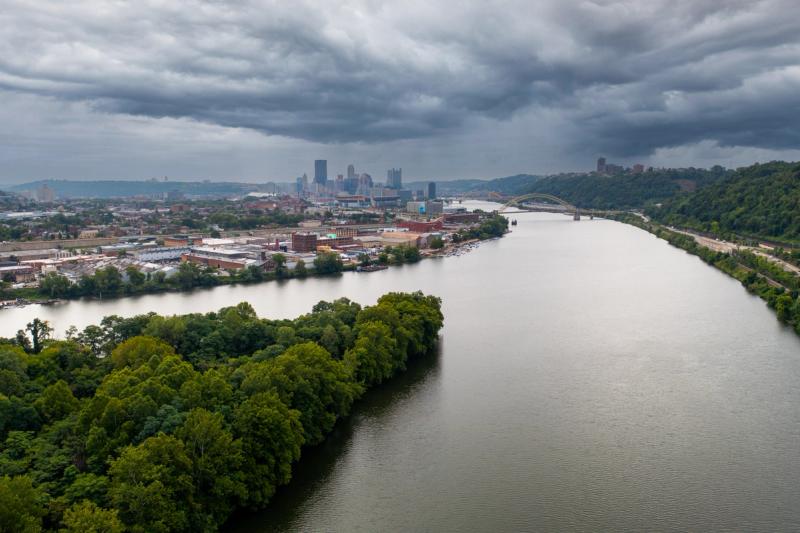 Aerial drone shot looking down a Pittsburgh river toward the distant downtown skyline, with dramatic dark storm clouds filling the sky above a green tree-lined bank.