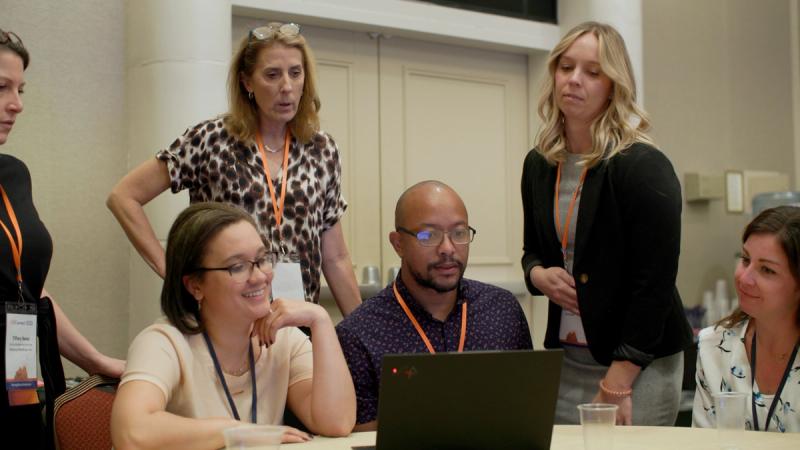 A group of five conference attendees huddles around a laptop at a workshop table, engaged in conversation and collaborative problem-solving.