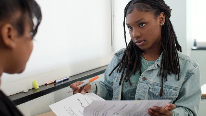Two high school students sit together reviewing a document, one pointing to the page while the other listens attentively.