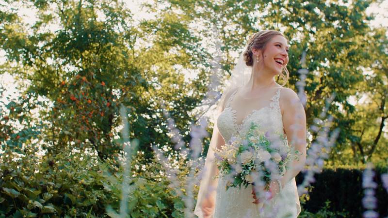 A bride in a lace gown and veil laughs joyfully while holding a bouquet of white and lavender flowers, standing in a lush garden with soft backlit foliage behind her.