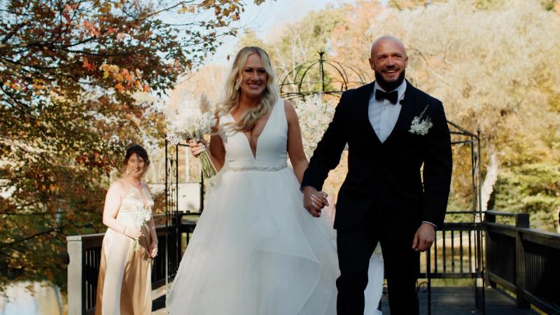 A bride and groom walk hand in hand through a wrought iron garden gate after their ceremony, beaming at guests, with autumn foliage and a bridesmaid visible behind them.