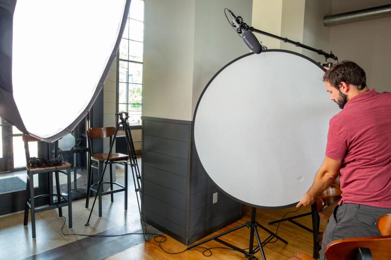 A Meridian Media crew member positions a large circular reflector during lighting setup for a video shoot, with a softbox and boom microphone visible in the background.
