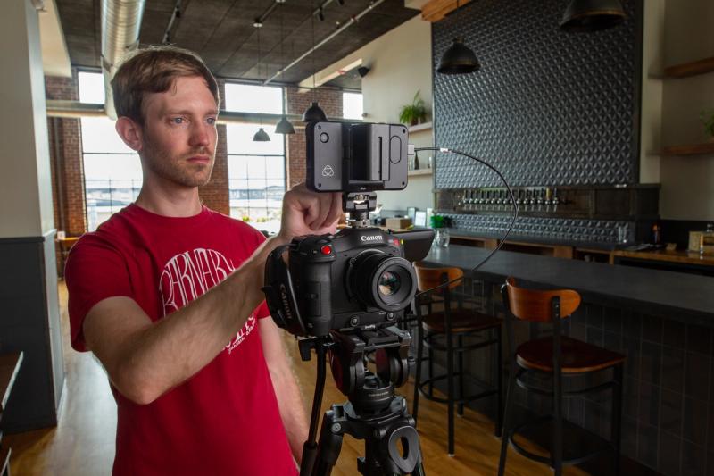 A Meridian Media videographer adjusts an external monitor on a Canon cinema camera mounted on a tripod, set up inside the Old Thunder Brewing Company taproom.