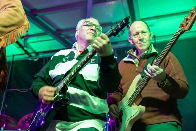 Two guitarists perform side by side on stage at Rock Against Cancer, lit by green stage lighting, with one focused intently on his instrument in the foreground.