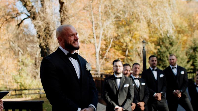 A groom in a black tuxedo smiles while standing at an outdoor autumn ceremony altar, with groomsmen in matching black tuxedos visible behind him against a backdrop of golden fall foliage.