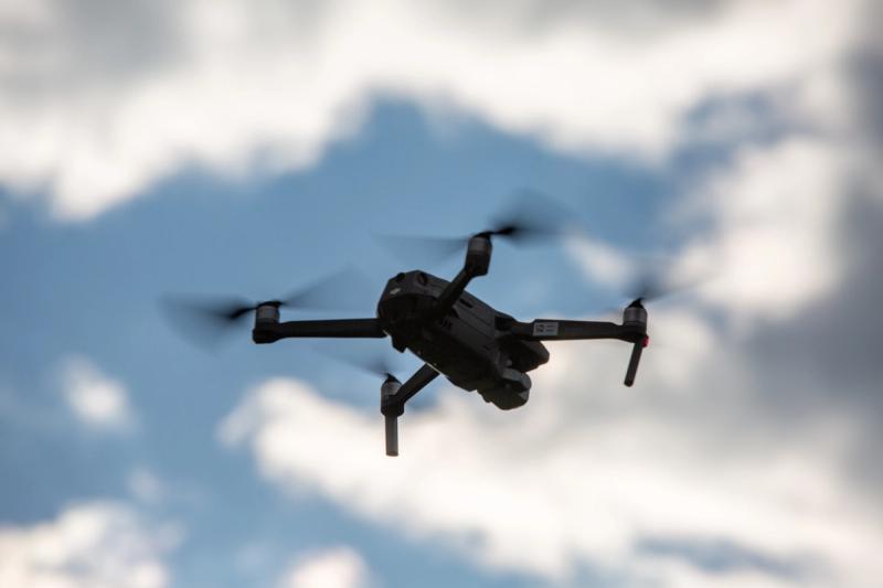 A DJI drone in flight against a partly cloudy blue sky, shot with a shallow depth of field that keeps the aircraft sharp against soft blurred clouds in the foreground and background.