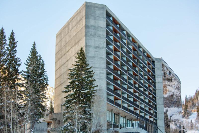 A modern hotel building at Snowbird resort in Utah, surrounded by pine trees with snow-covered mountains visible in the background under a clear winter sky.