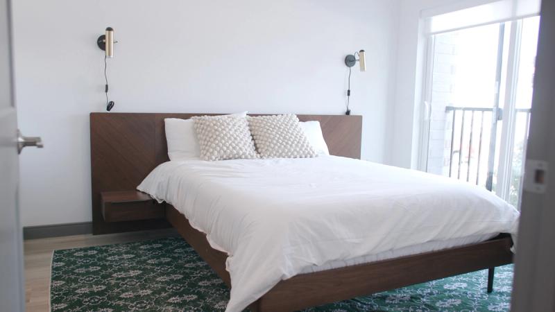 A bright model bedroom at Brewer's Block with a walnut platform bed, white bedding, wall-mounted reading lights, a green patterned area rug, and a glass balcony door letting in natural light.