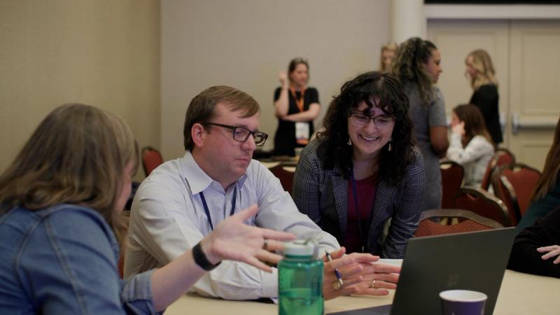 Two conference attendees sit at a workshop table in conversation, one gesturing expressively while the other listens and smiles, with other participants visible in the background.