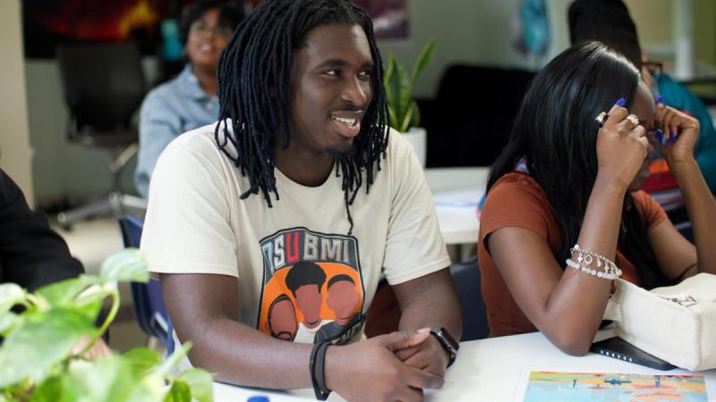 A male high school student with blue locs smiles and looks to the side while seated at a table in a classroom, with other students visible around him.