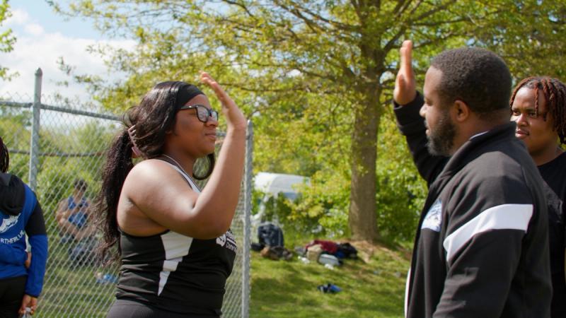 A high school student in a track uniform high-fives a coach outdoors at a track meet, with other students visible in the background.