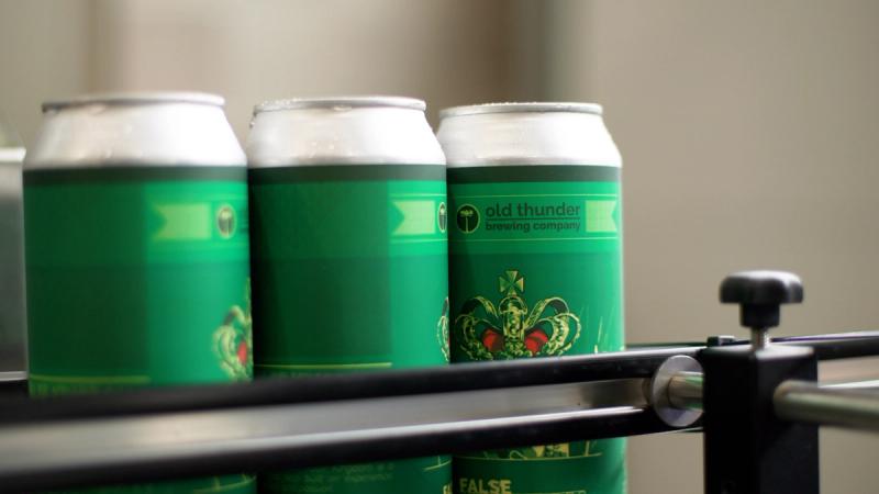 Three labeled Old Thunder Brewing Company cans of False Kingdom IPA on a canning line, showing the green label design with a crown illustration.