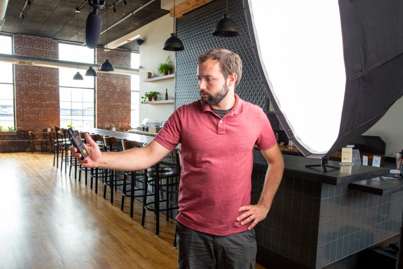 A videographer holds a light meter while standing in the taproom at Old Thunder Brewing Company in Pittsburgh, with a large round softbox visible to the right and the brewery's bar and windows in the background.