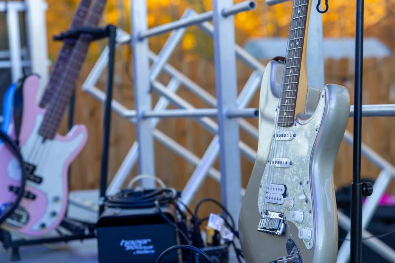Two electric guitars on stands backstage before the Rock Against Cancer performance, with autumn foliage visible in the background and a pink bass guitar and amplifier visible out of focus.