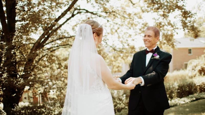 A bride in a lace wedding gown and veil holds hands with her father outdoors during a first look moment, surrounded by autumn trees with warm golden light.