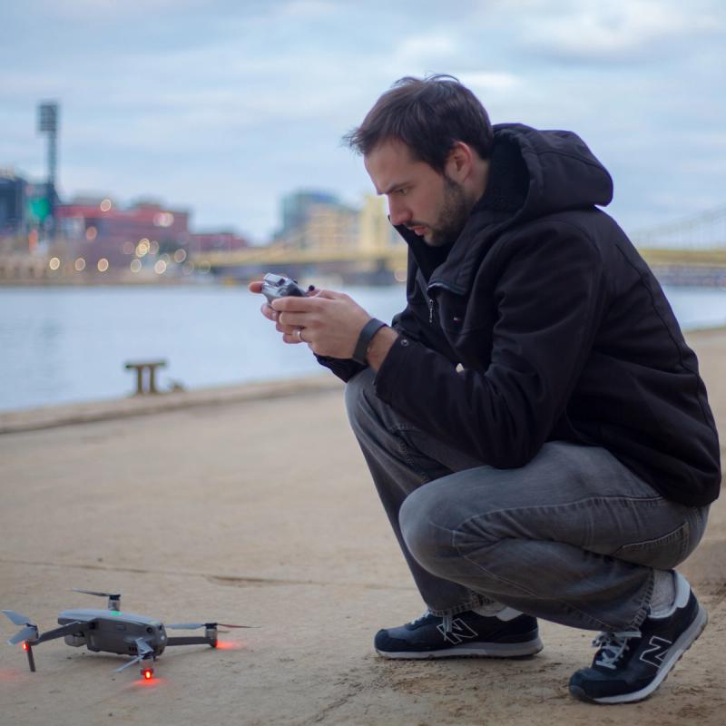 A videographer crouches on a riverfront path in Pittsburgh preparing to launch a DJI drone, with the controller in hand, the North Shore bridges and river visible in the soft-focus background.