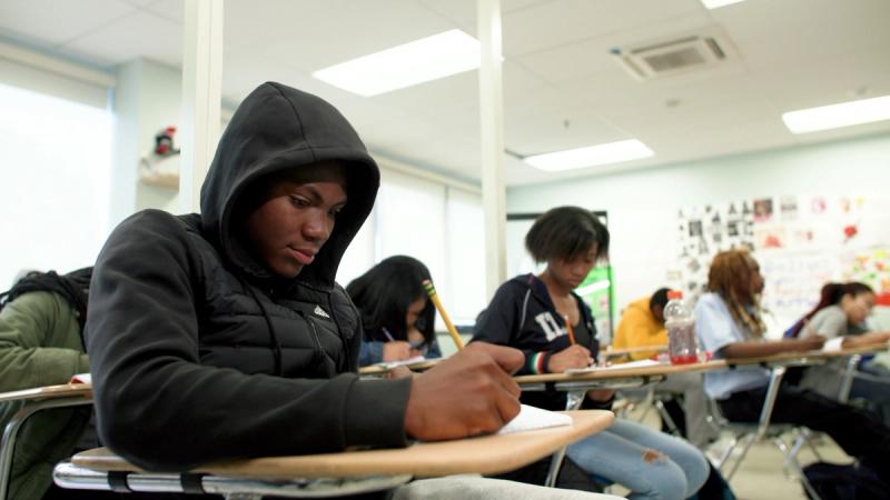 A high school student in a black hoodie writes at a desk during class, with other students visible working in the background at Freire Charter School Wilmington.