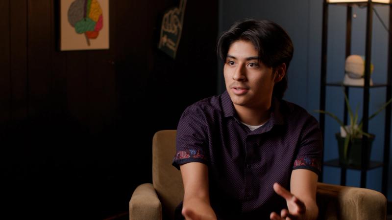 A high school student sits in a warmly lit interview setting and speaks expressively, with classroom posters and a plant visible in the background.