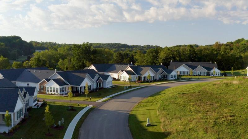 Low-angle drone shot of a row of completed Weaver Homes at Heritage Crossings, with a gently curving road, manicured lawns, and a treeline visible in the background under a partly cloudy sky.