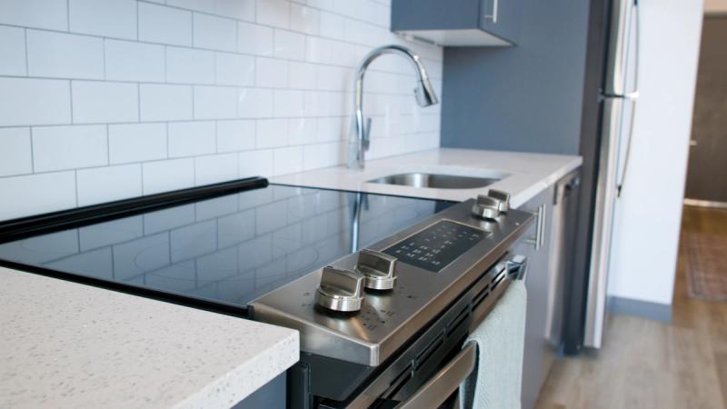 A modern apartment kitchen at Brewer's Block featuring a black glass electric range, quartz countertops, white subway tile backsplash, blue-gray cabinetry, and stainless steel appliances.
