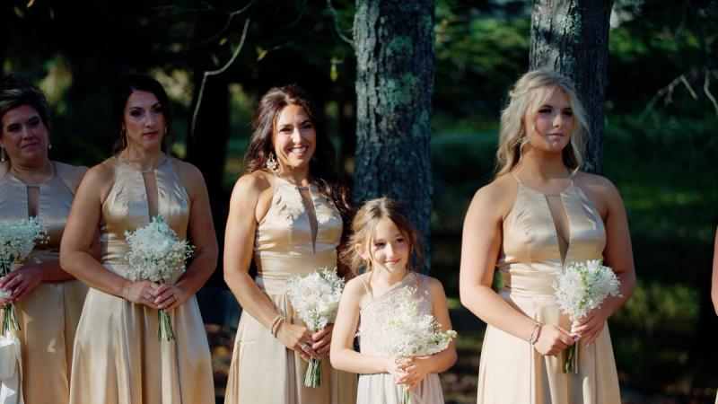 A group of bridesmaids in champagne gold gowns holding white bouquets stand outdoors among tall pine trees, with a young flower girl in a white dress smiling in the center.
