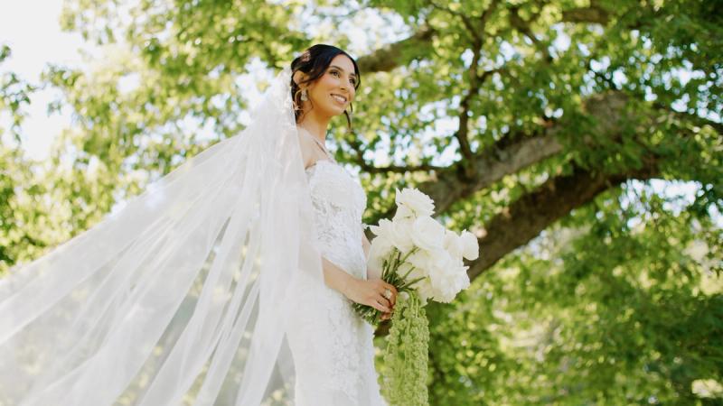 A bride in a lace off-shoulder wedding gown holds a white bouquet and smiles in a sun-dappled garden at Springton Manor Farm, her veil caught mid-movement by a breeze.
