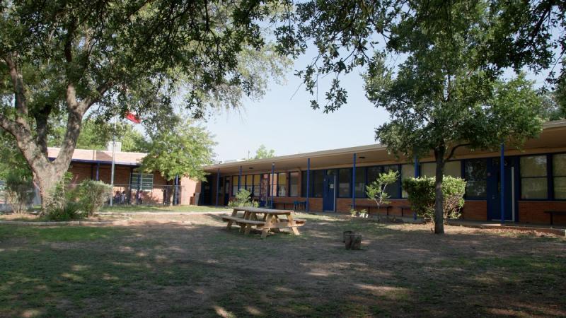 The exterior of a single-story brick elementary school in Austin, Texas, with large oak trees providing shade over a courtyard with picnic tables on a sunny day.