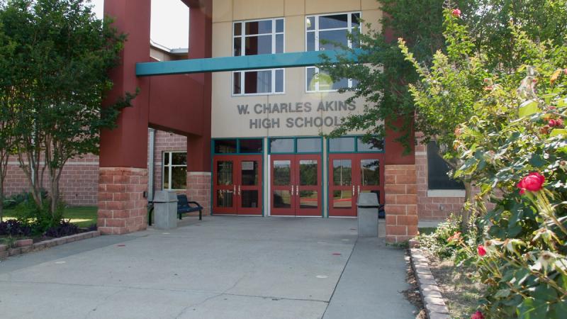 The front entrance of W. Charles Akins High School in Austin, Texas, with red brick walls, teal accent details, red entry doors, and flowering shrubs in the foreground.