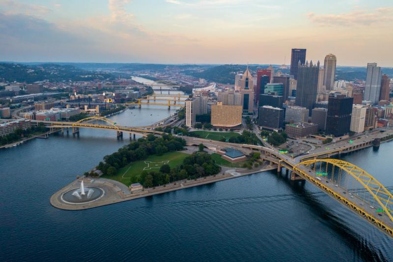 Aerial drone shot of downtown Pittsburgh from above the Point, showing the Fort Pitt Bridge, Point State Park fountain, the confluence of the rivers, and the full downtown skyline under an evening sky.