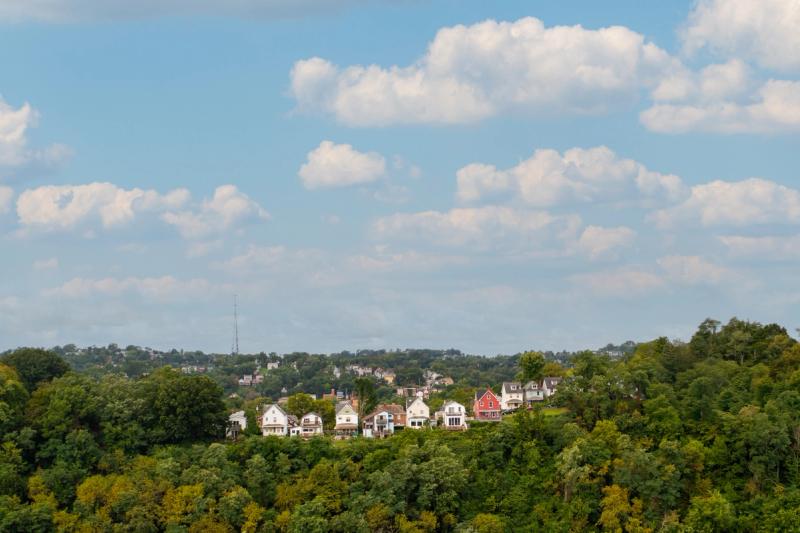 Aerial drone shot of a Pittsburgh hillside neighborhood with colorful houses emerging from a dense tree canopy under a partly cloudy blue sky.