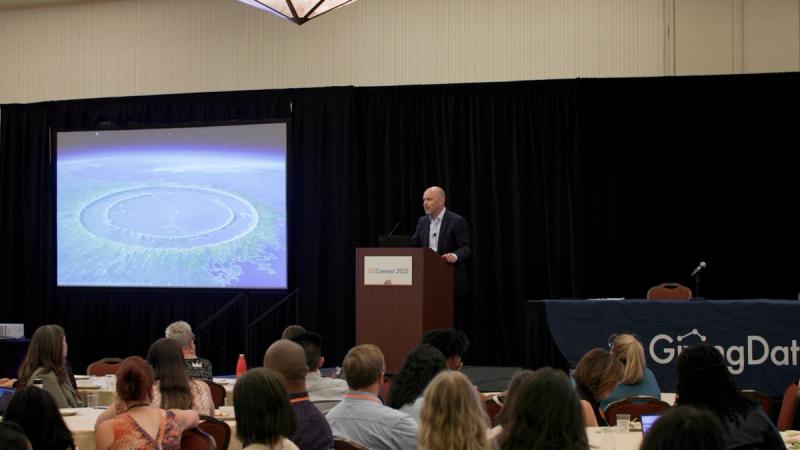 A speaker presents at the GivingData Connect conference, standing at a podium in front of a large projected image of Earth from space, with a full ballroom audience seated at round tables.