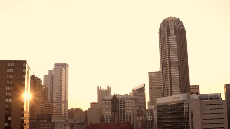 The Pittsburgh downtown skyline at golden hour, with warm amber light flooding between the skyscrapers and the sun visible as a starburst between two buildings.