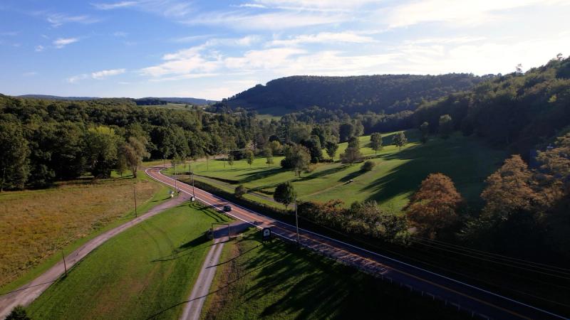 Aerial drone shot of a rural two-lane road winding through rolling green hills and forested ridges near Chanteclaire Farm in Friendsville, Maryland, under a blue sky with light clouds.