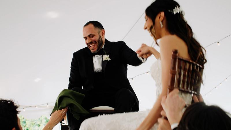 A groom is lifted in a chair during the Hora at an outdoor tented reception at Springton Manor Farm, laughing as guests celebrate around him, with string lights visible overhead.