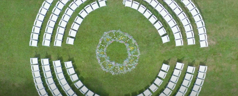 Aerial drone shot looking straight down at a circular outdoor wedding ceremony setup on a green lawn, with white chairs arranged in concentric arcs around a floral wreath centerpiece.