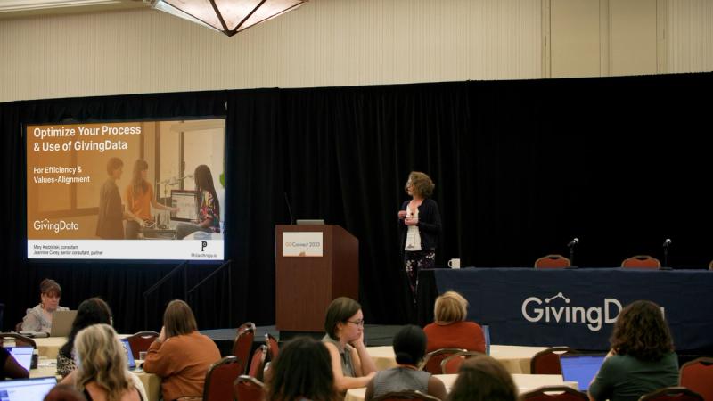 A presenter stands at a podium at GivingData Connect, gesturing toward a projected slide titled "Optimize Your Process & Use of GivingData — For Efficiency & Values-Alignment."