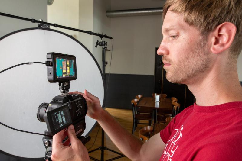 A videographer adjusts a camera monitor during a video interview setup, with lighting equipment visible in the background.
