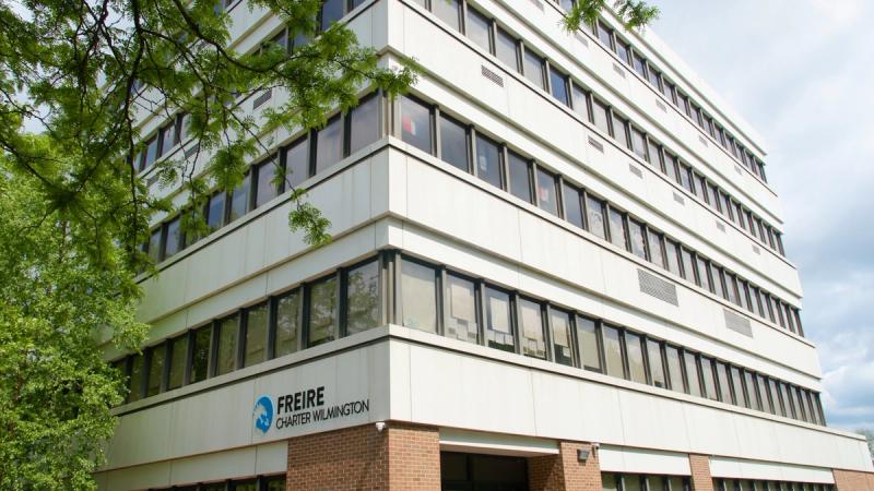 he exterior of the Freire Charter School Wilmington building, a multi-story white structure with large windows and the school's logo visible at street level, surrounded by trees.