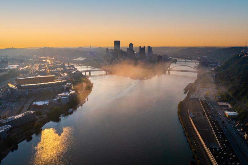 erial drone shot of Pittsburgh at sunrise, with golden light and morning mist over the Ohio River, the downtown skyline visible in the distance and Acrisure Stadium on the left.